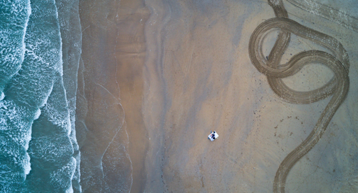 Aerial view of a beach with visible waves and trails in the sand left by some kind of truck or bulldozer